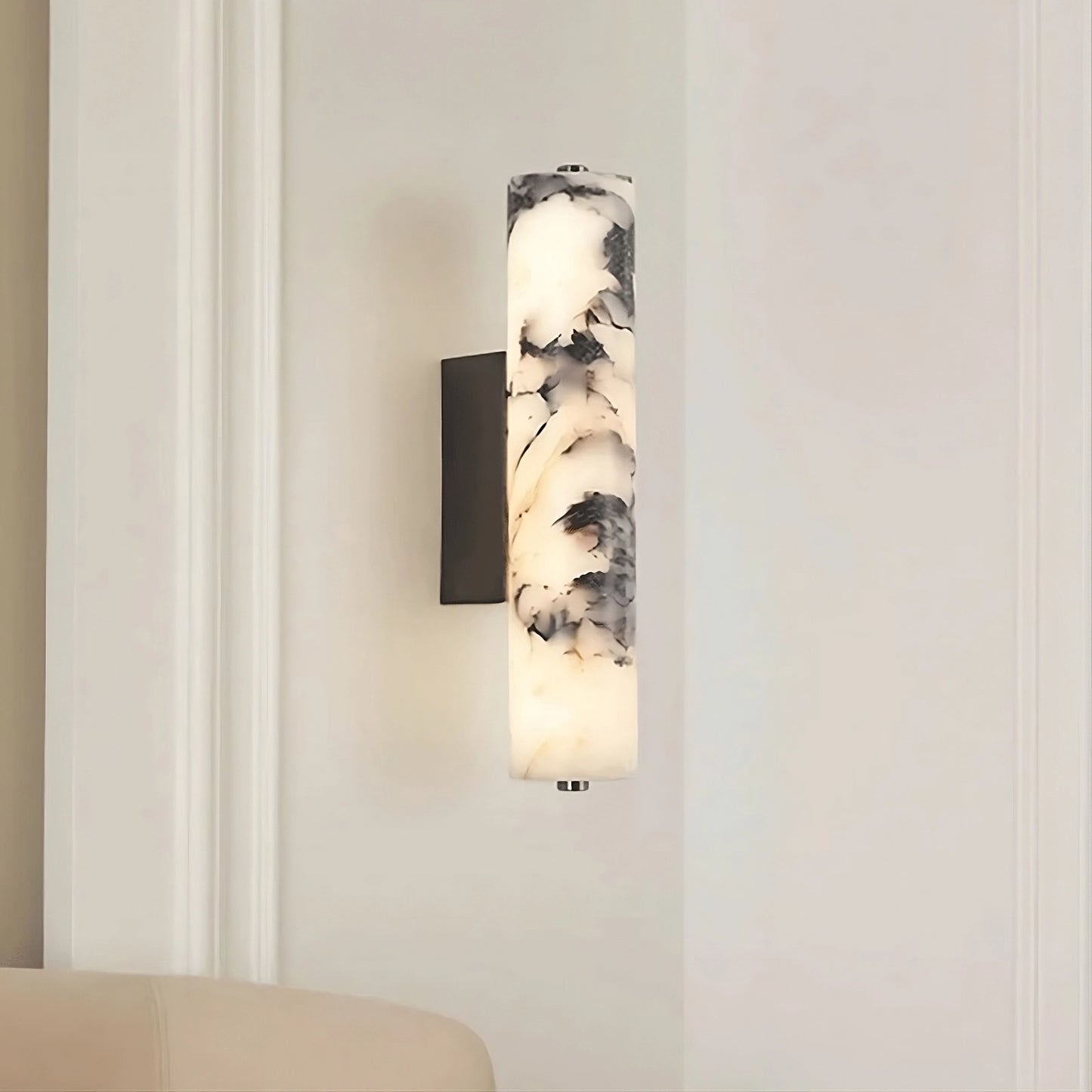 The Valmont Marble Cylinder Sconce by Morsale features a cylindrical marble-patterned shade and emits soft light. It is mounted on a light-colored wall above a beige upholstered chair.