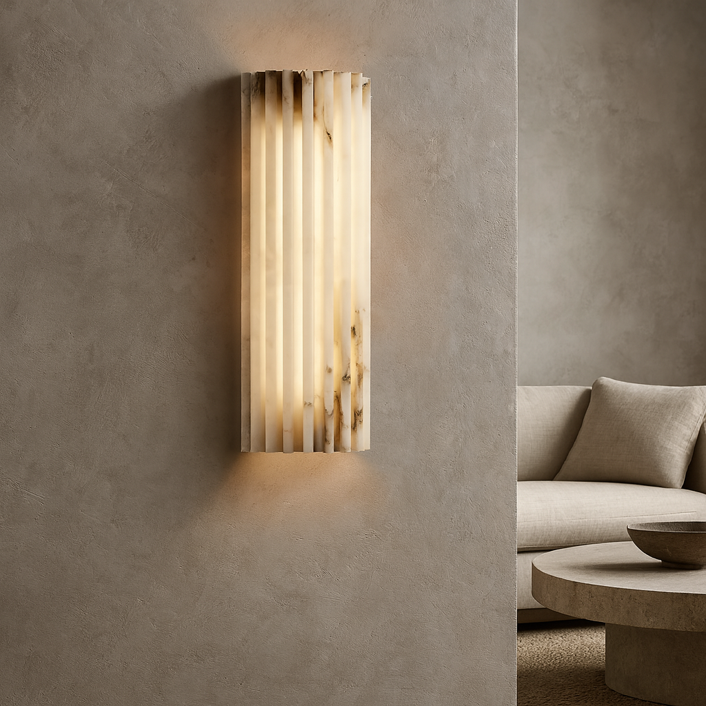 The Marlo Marble Wall Sconce 12" by Morsale casts a warm glow on a textured beige wall. In the background, a minimalist living room includes a beige sofa and round coffee table.