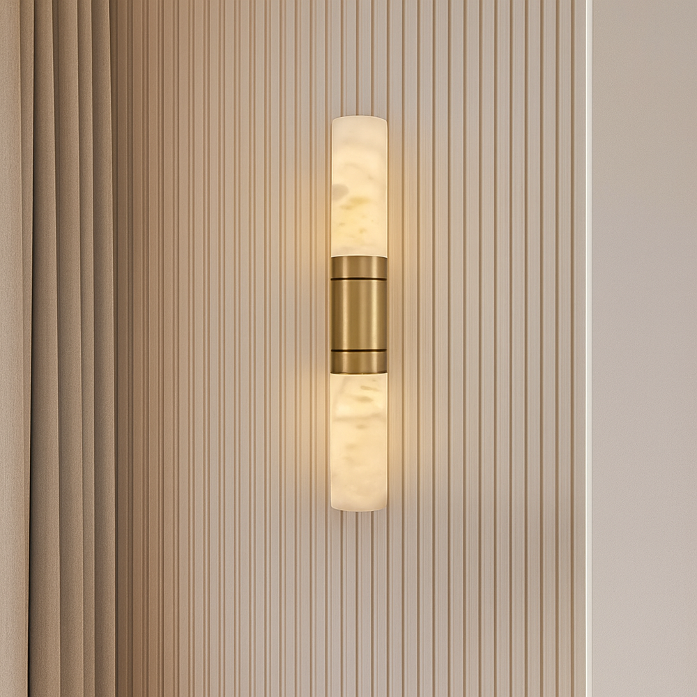 The Solenne Marble & Brass Wall Sconce by Morsale.com features a luxurious brass center and frosted glass ends, casting a warm glow against a vertically grooved beige wall beside matching beige drapery.