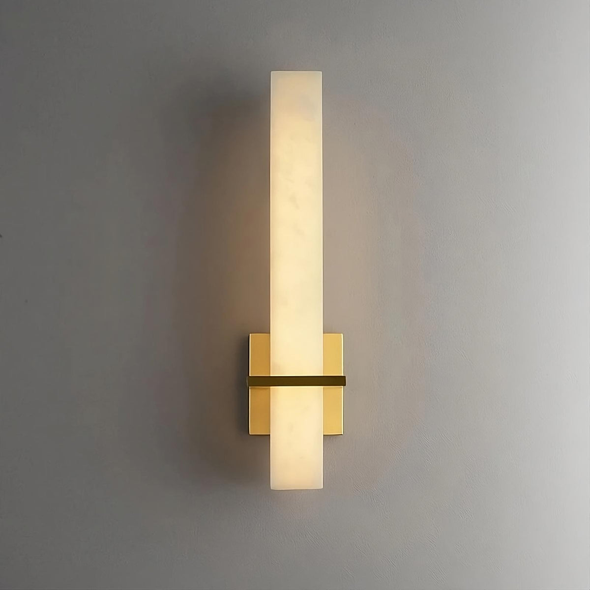 A modern wall-mounted brass light fixture with a vertical rectangular ivory-colored shade and a gold base is illuminated against a plain gray background. The Natural Marble Indoor Sconce by Morsale.com has a minimalist design, casts a soft, warm light, and exemplifies the elegance of LED indoor lighting.