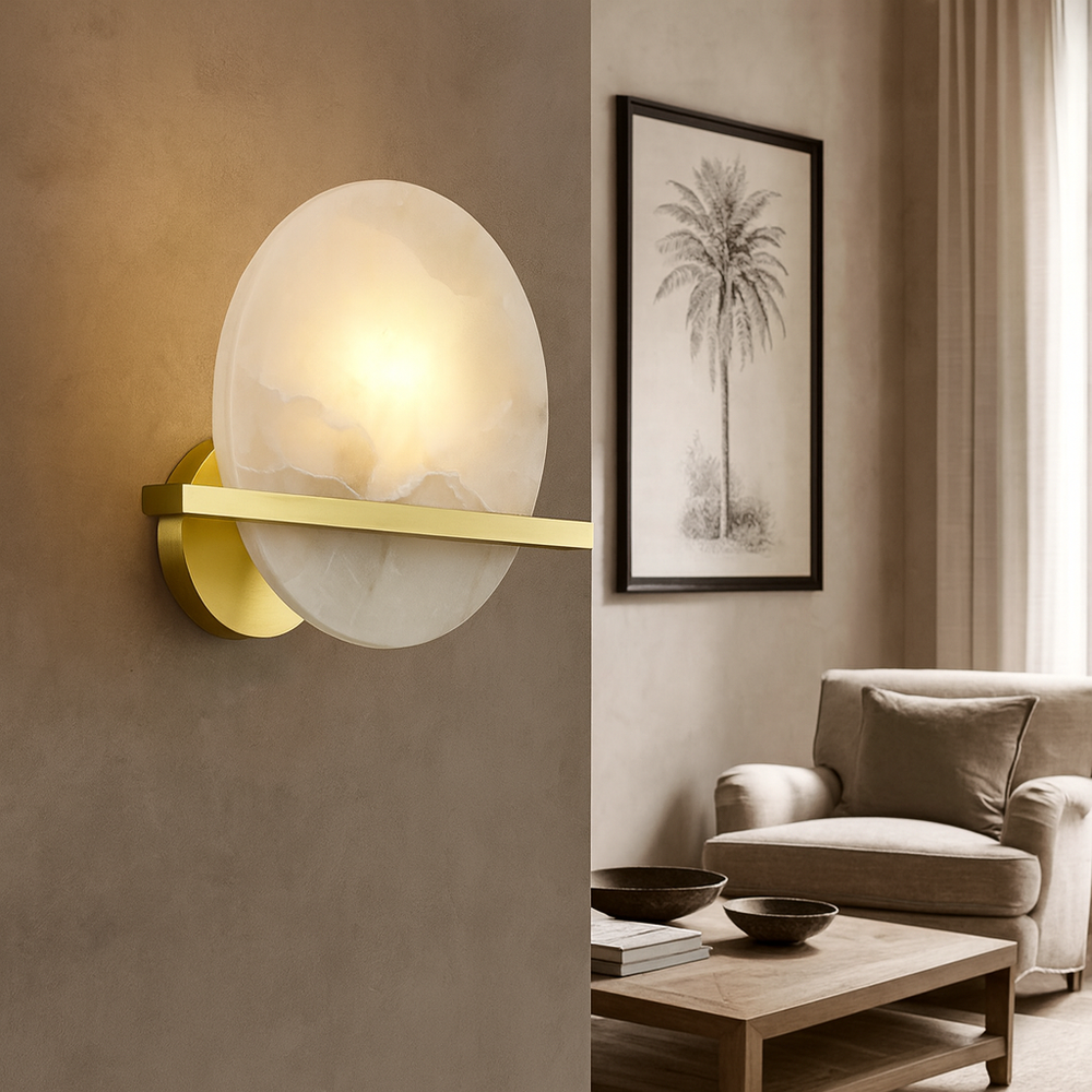 
                  
                    The Elara Natural Marble Wall Sconce by Morsale.com features a round, frosted glass shade and brass details, softly illuminating a stylish living room with neutral décor, framed palm tree art, and cozy seating.
                  
                