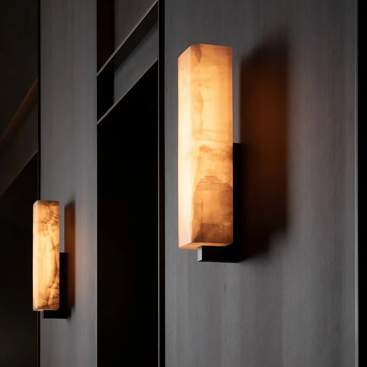 Mounted on a sleek, dark wall, two Morsale Alton Marble Wall Lamps in warm amber emit a soft LED glow with their modern rectangular design and marbled pattern, creating a cozy ambiance.