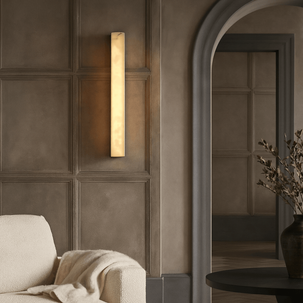 The Linden Marble Sconce from Morsale.com softly illuminates a beige paneled wall beside a cozy cream sofa with a blanket, near an arched doorway and a dark vase filled with green branches.