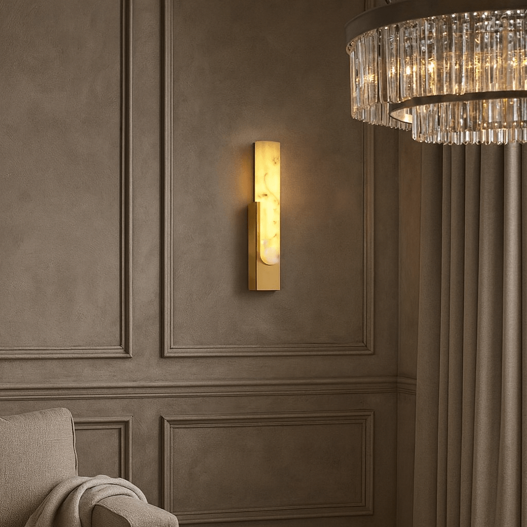 The Wexford Marble Wall Sconce by Morsale.com glows on a taupe paneled wall beside beige curtains, a crystal chandelier, and a light-colored upholstered armchair with a draped blanket.
