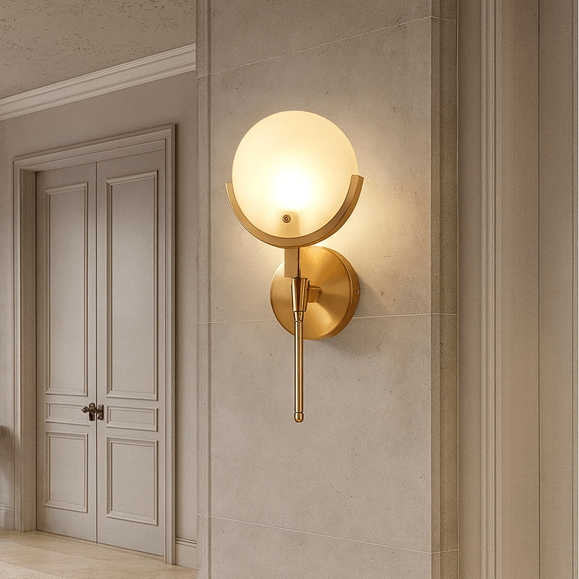 The Vellaro Marble Wall Sconce from Morsale.com, featuring a round frosted glass globe and gold accents, is mounted on a beige stone wall in an elegant hallway with double doors, a rug, and a decorative branch on a small table.