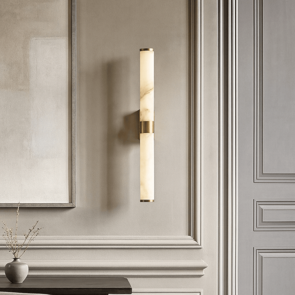 
                  
                    The Morsale Altura 27" Marble & Copper Sconce, featuring a cylindrical marble-like shade and copper accents, is mounted on a neutral paneled wall near a minimalist table with a vase of dried branches.
                  
                