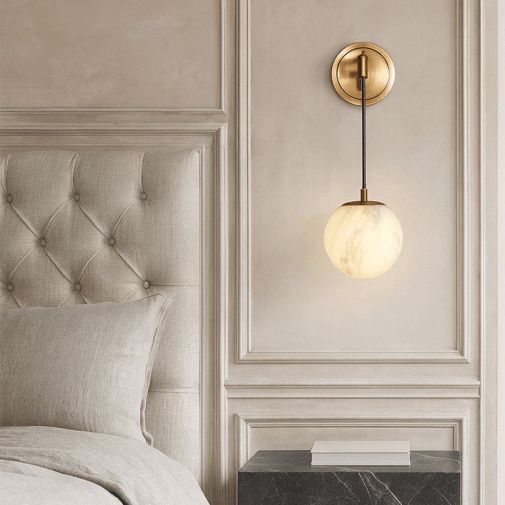 A modern bedroom corner featuring a tufted beige headboard, neutral bedding, a black marble nightstand, and the Morsale Horrin Marble Ball Wall Sconce with a glowing glass globe on an ornate cream wall.