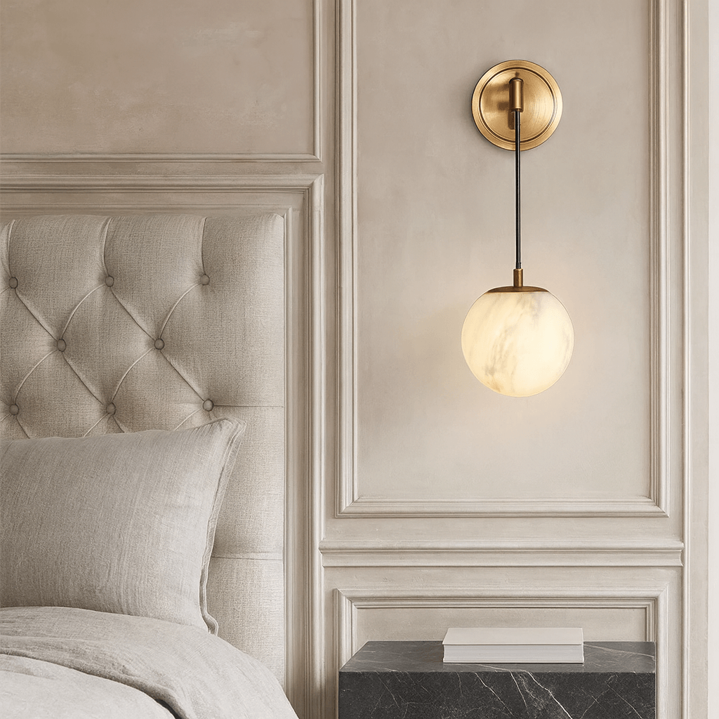 A modern bedroom corner featuring a tufted beige headboard, neutral bedding, a black marble nightstand, and the Morsale Horrin Marble Ball Wall Sconce with a glowing glass globe on an ornate cream wall.