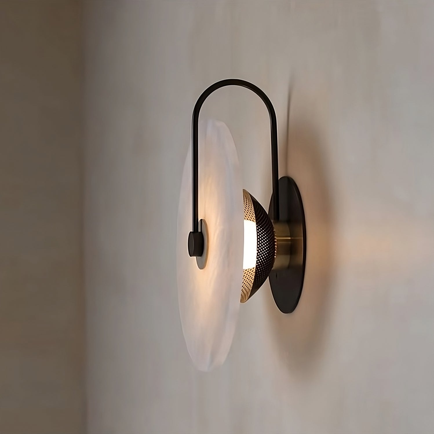 The Moonshade Natural Marble Sconce Light by Morsale.com offers a modern design with its circular, translucent diffuser and black metal frame, emitting a warm glow against a neutral wall. Equipped with LED chips and mesh detailing around the light source, it brings a contemporary flair to any space.