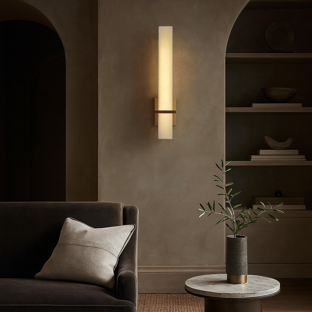 A modern living room with a dark sofa, light pillow, and round table with a vase of leafy branches features the Varella Natural Marble Wall Sconce by Morsale.com glowing warmly against a textured beige wall with built-in shelves.
