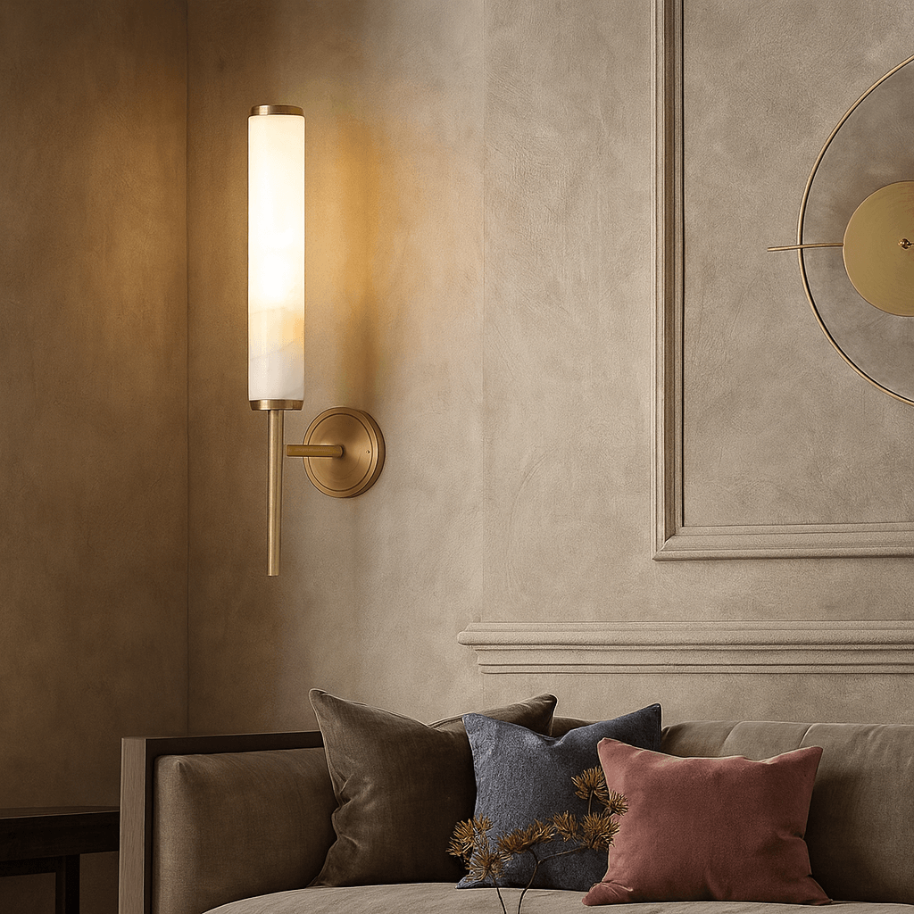 The Burston Marble Wall Sconce by Morsale.com, featuring a cylindrical shade, is mounted above a beige sofa with throw pillows, set against a textured light wall with decorative molding.