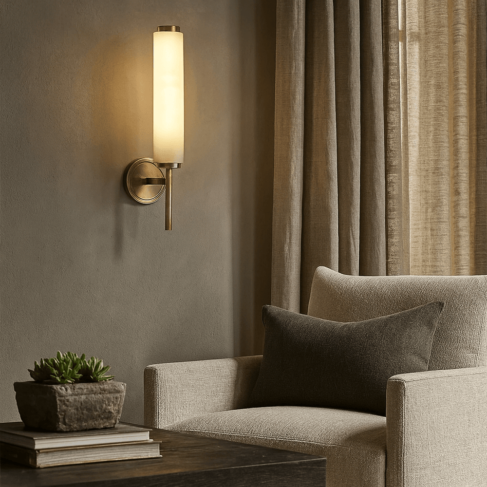 
                  
                    The Burston Marble Wall Sconce by Morsale.com, featuring a cylindrical frosted glass shade, glows above a beige armchair with a dark pillow beside a window and wooden side table topped with books and a potted plant.
                  
                