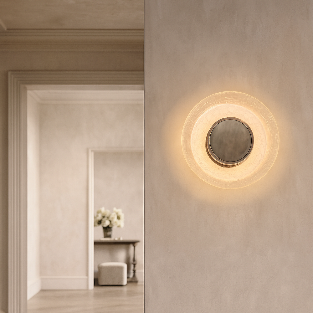 The Celeste Dish Sconce by Morsale features a modern circular marble design with a glowing light around a central mirror, mounted on a beige wall. In the background, a doorway opens to a softly lit room with a table and white flowers.