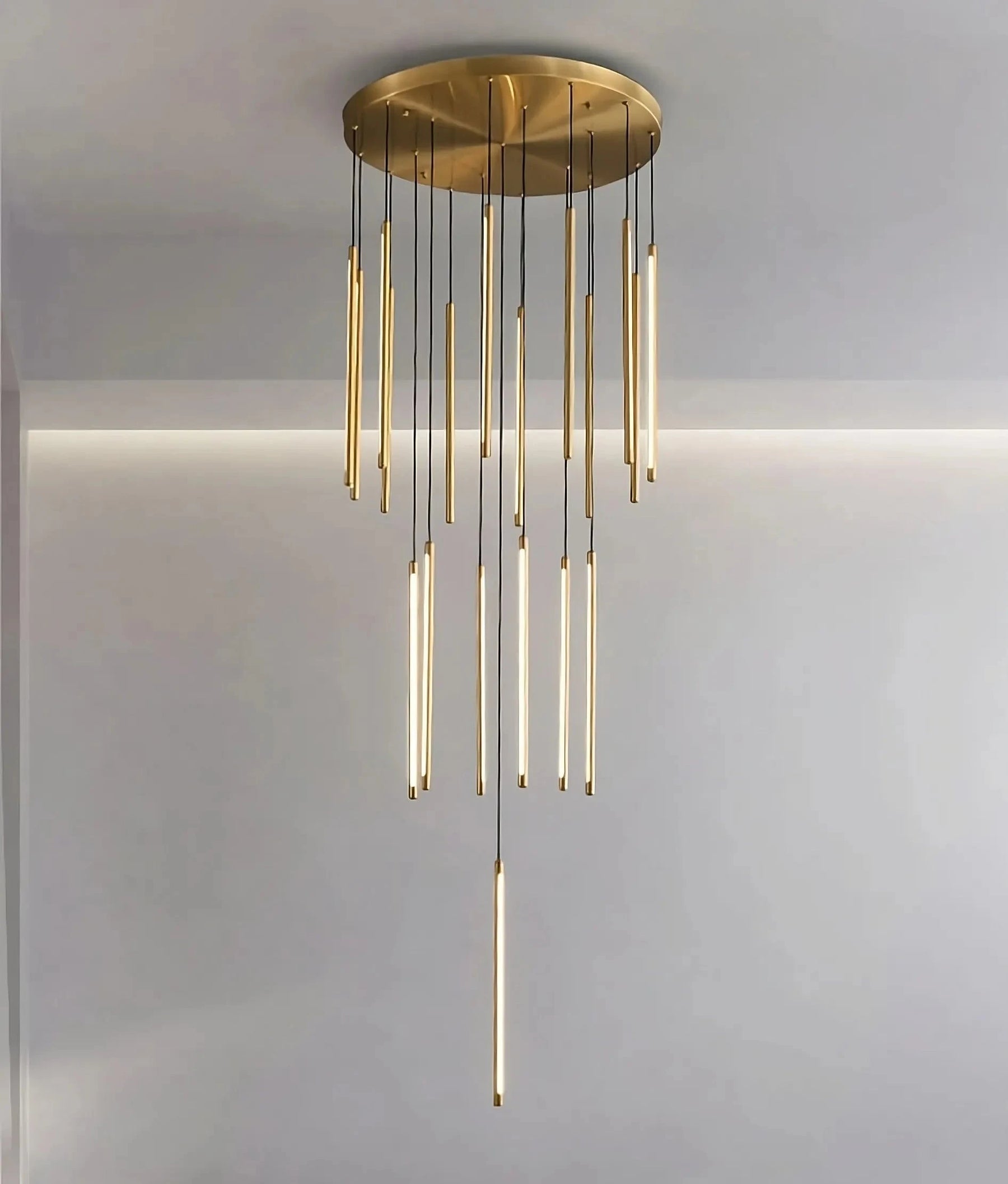 The Grande Brass Entryway Pendant Light Fixture from Morsale.com is an elegant, modern chandelier with a golden circular base and multiple long, slender vertical light rods hanging at varying lengths. It features dimmable LED chips that cast a soft, warm glow against a minimalistic white ceiling and wall background.