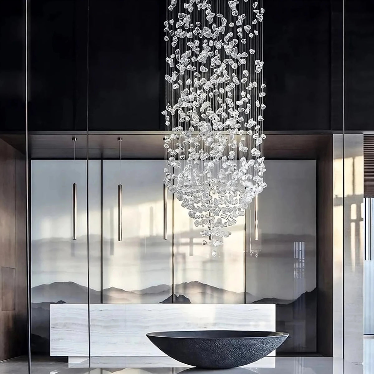 A modern interior is illuminated by the elegant Multi Crystal Pendant Duplex Chandelier from Morsale.com, a large chandelier adorned with cascading crystal accents. Below, a decorative, wide, black basin rests on a white countertop. The background showcases a mural of misty mountains and sleek vertical pendant lights.