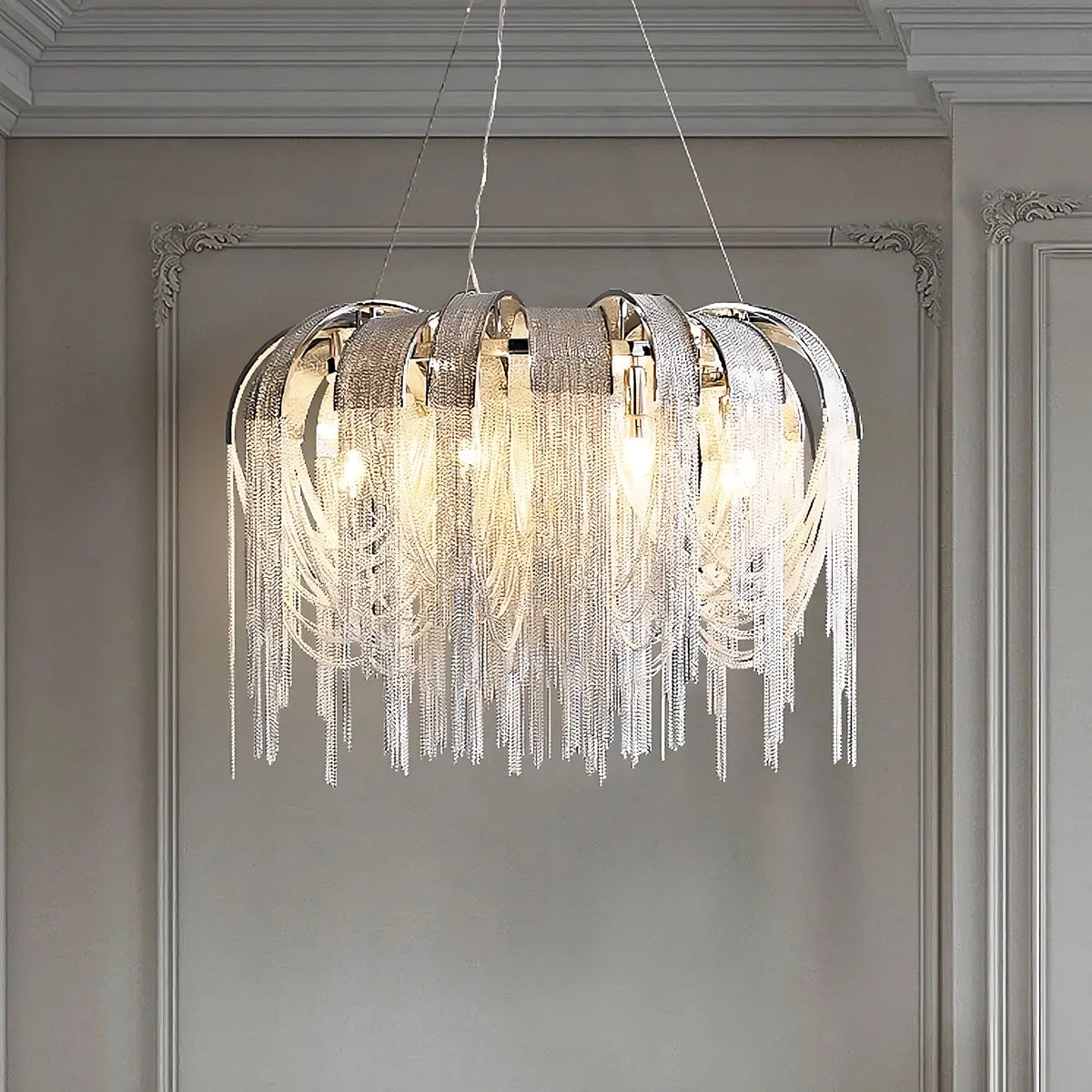 A sophisticated Cadena Contemporary Chandelier from Morsale.com, with a modern design, boasts polished silver curved arms and cascading strands of fine, shimmering crystals. Hanging from the ceiling with its adjustable wire and equipped with LED bulbs, it emits a warm glow that illuminates the ornate room adorned with intricate molding details.