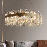 The Asti Crystal Modern Chandelier from Morsale.com, featuring a luxurious stainless steel frame and an array of handmade crystal prisms, is suspended from the ceiling. Emitting a warm, bright light, it illuminates a beige wall and a portion of a couch with cushions in the foreground.