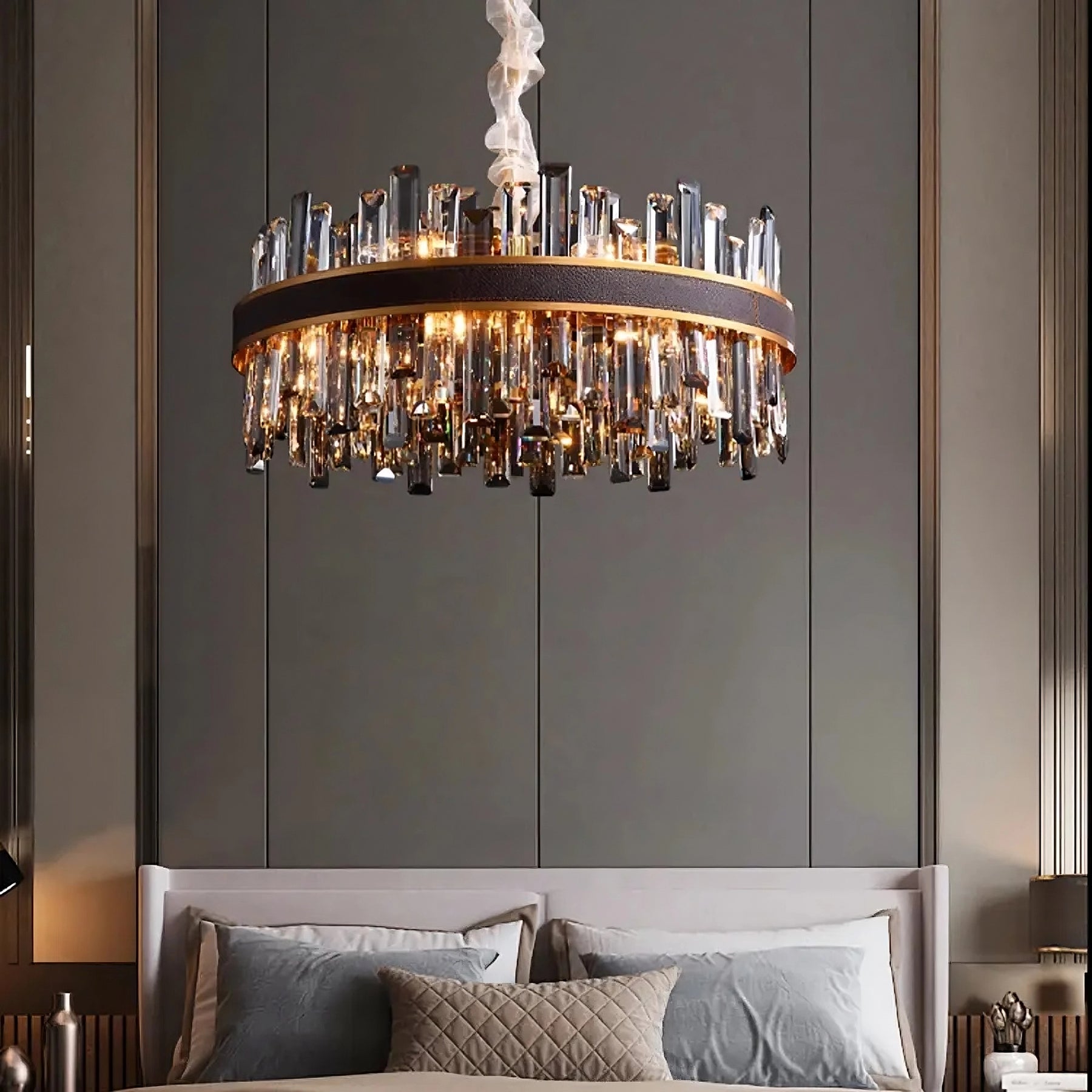 A luxurious bedroom features a Calvi Modern Chandelier by Morsale.com hanging from the ceiling, centered above a neatly made bed with light gray bedding. The chandelier has a circular, gold-plated stainless steel frame adorned with handmade crystals, creating an elegant and sophisticated atmosphere.