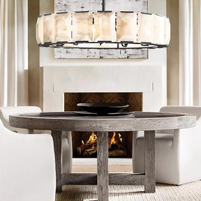 A cozy dining room featuring a round wooden table with a black bowl centerpiece, under an elegant Villano Calcite Crystal Chandelier from morsale.com. A lit fireplace adds warmth to the serene setting.