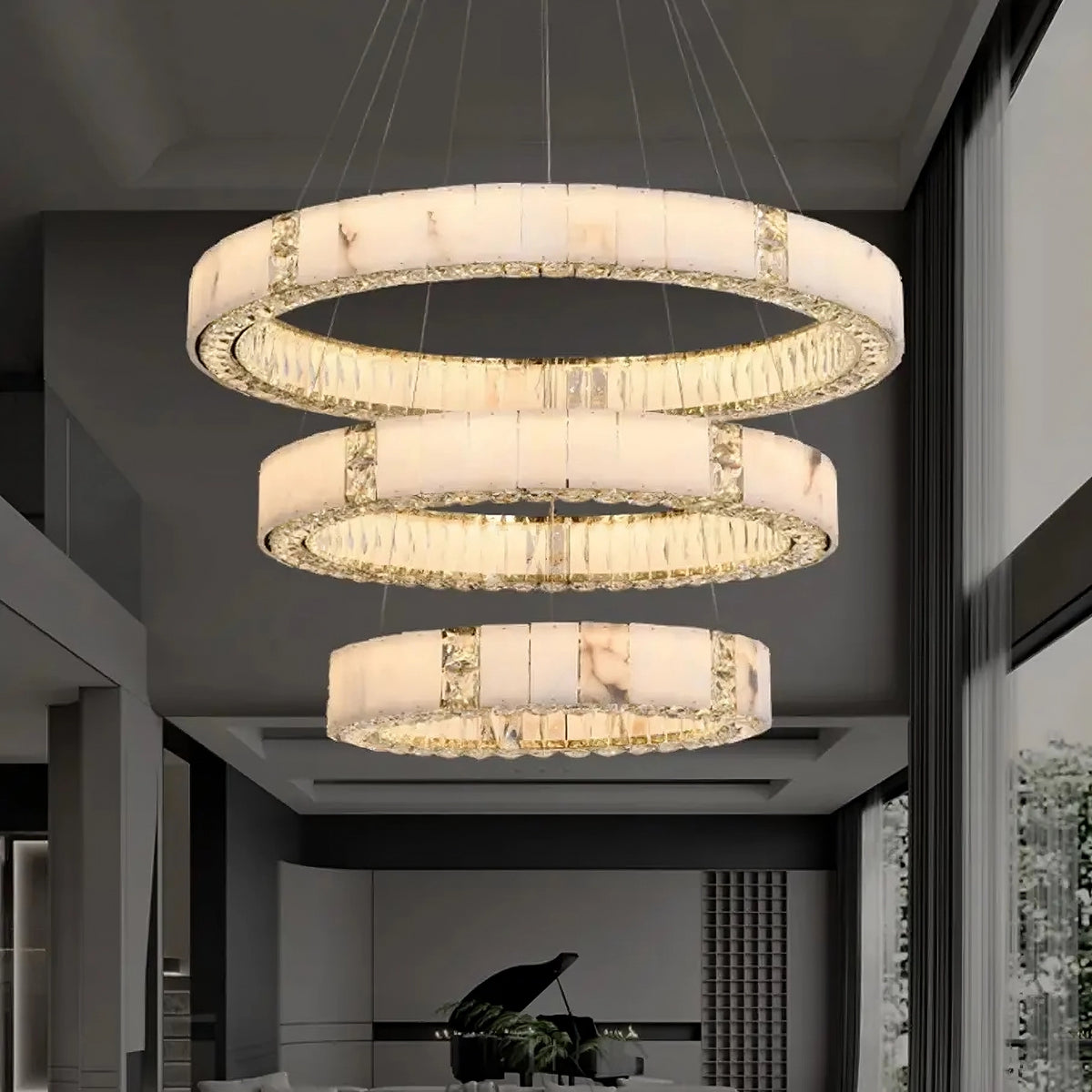 A modern indoor space features a striking Marble & Crystal Modern Chandelier from Morsale.com with circular, translucent rings. Each ring is illuminated, casting a soft glow. Tall windows allow natural light to enter, and a grand piano is partially visible in the background.