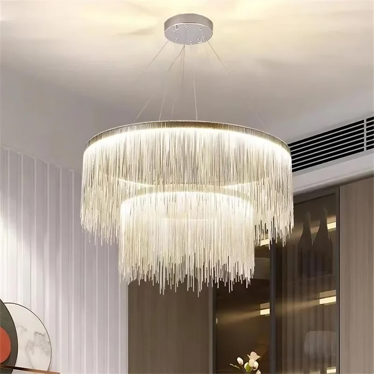 The Cadena Steel Tassel Modern Chandelier by Morsale.com, featuring multiple layers of cascading thin golden chains in a large circular design, serves as the focal point with its sleek LED light source. The backdrop of marble walls with a white and gray veined pattern underscores the elegance of the modern interior design.