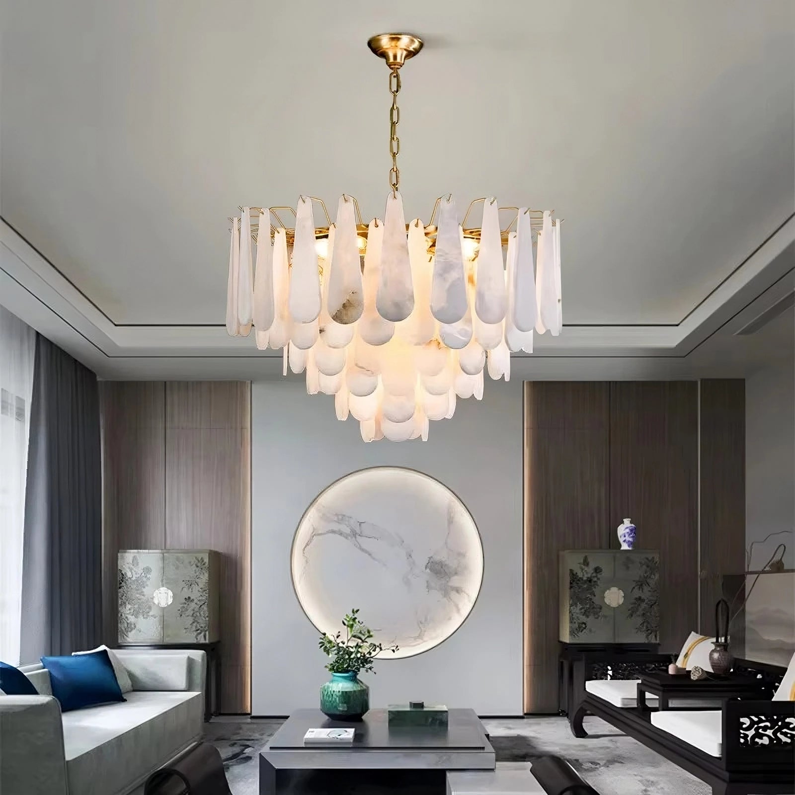 A modern living room showcases the luxurious Moonshade Marble Chandelier by Morsale, featuring white, petal-like pendants and energy-efficient LED bulbs. The space includes a gray sofa, round marble artwork adorning the wall, a coffee table adorned with a potted plant, and large windows inviting natural light to enhance the perfect modern style.
