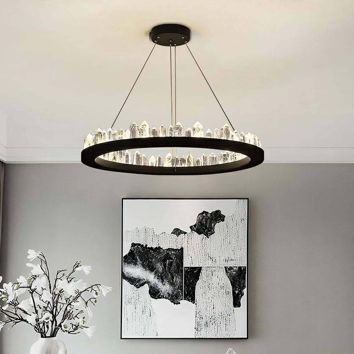 A Villa Collection Crystal Wheel Chandelier from Morsale.com hanging above a monochrome abstract painting on a wall, with a vase of white flowers on the left—elegant interior setting.