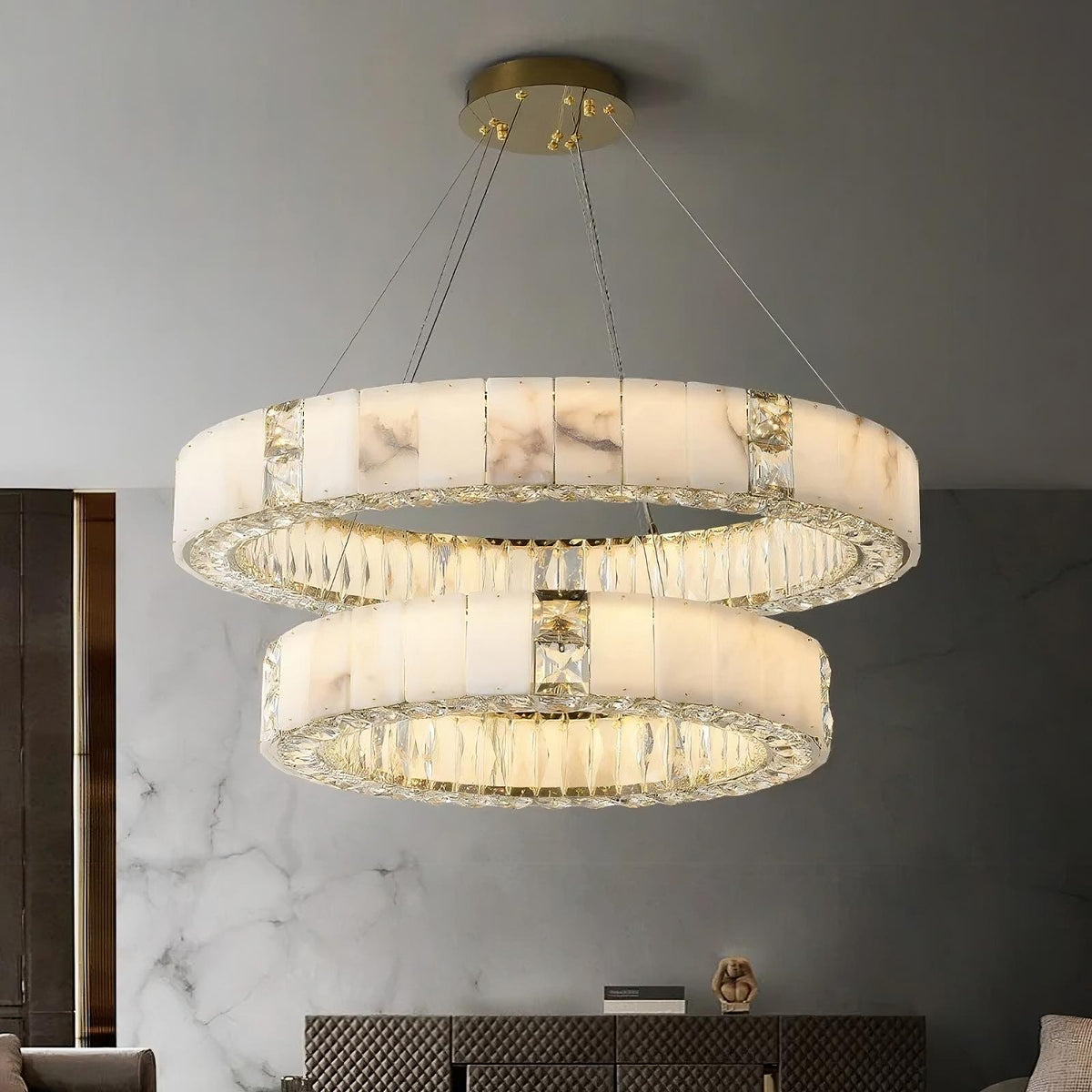 A modern ceiling chandelier, the Natural Marble & Crystal Modern Ceiling Light Fixture by Morsale.com, hangs elegantly, featuring two circular, marble-like rings with glass elements. Each ring is suspended by metal wires against a gray wall with a subtle marble pattern and a touch of luxury home décor visible below.