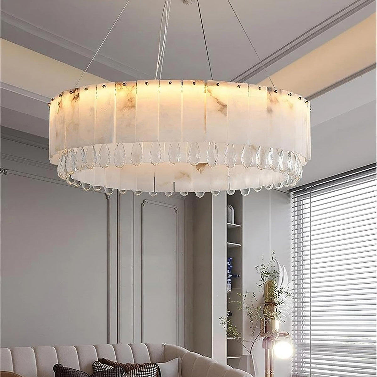 A modern living room features an elegant, large Modern Crystal and Spanish Marble Chandelier by Morsale.com with a circular design hanging from the ceiling. Below the chandelier is a taupe sofa adorned with pillows. Natural light filters in through a window with horizontal blinds. A decorative plant is visible.