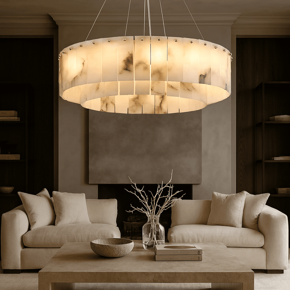 A modern living room features two cream sofas, a wooden coffee table with books and a vase of dried branches, and the Bigman Buckley Marble Modern Chandelier hanging above. Shelves flank a minimalist fireplace.