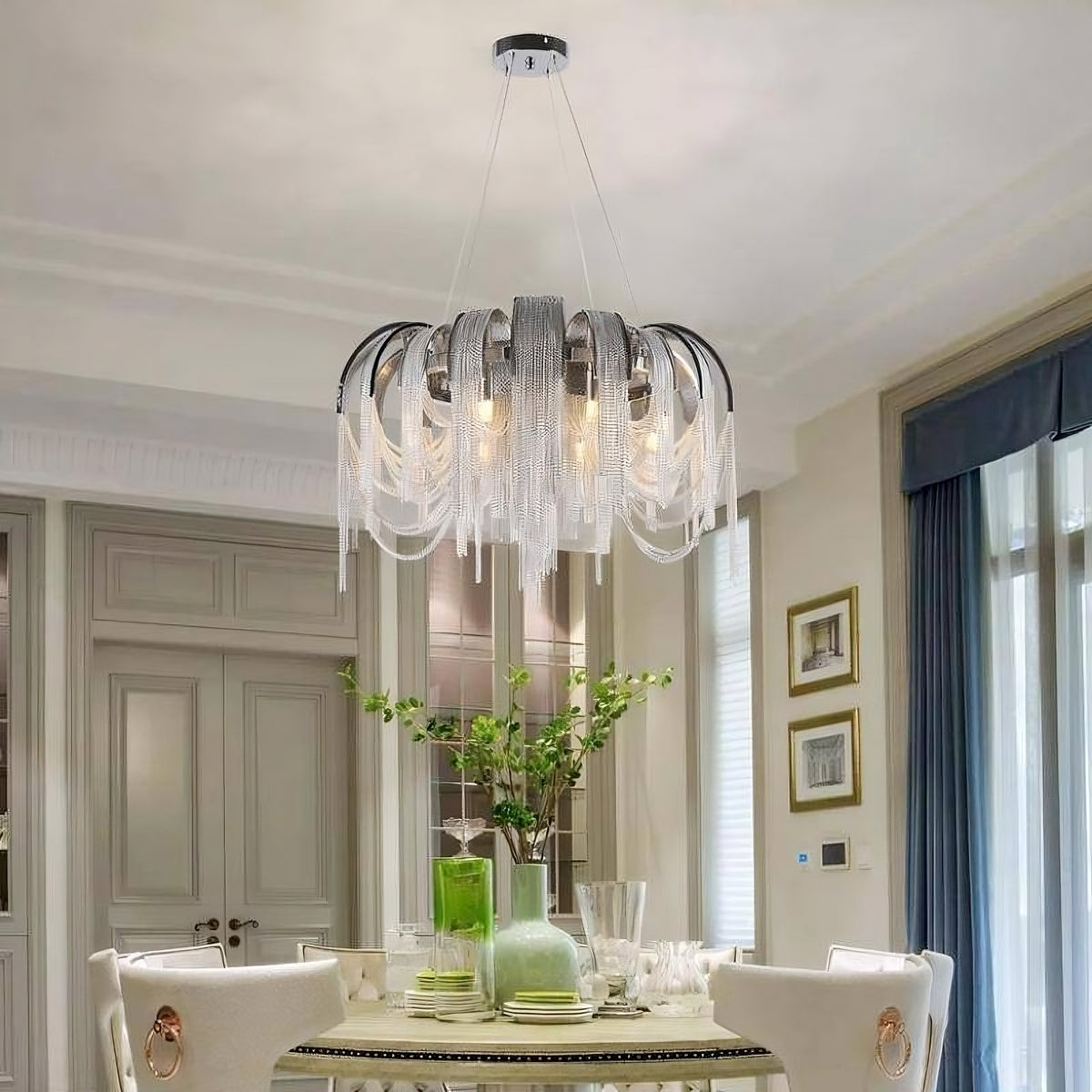 A dining room features a round table with elegant white chairs, a centerpiece with green plants, and glassware. Overhead is a Cadena Chain Tassel Modern Chandelier from Morsale.com with adjustable ceiling wire length and E14 LED bulbs. The room has large windows with blue curtains and framed artwork on the wall.