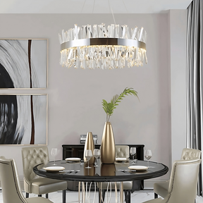 A modern dining room featuring a round black table set with wine glasses, dishes, and a vase with greenery. Four beige upholstered chairs surround the table. Above hangs an elegant Morsale.com Calvi Crystal Modern Chandelier with handmade clear crystals. A monochromatic abstract painting adorns the wall.