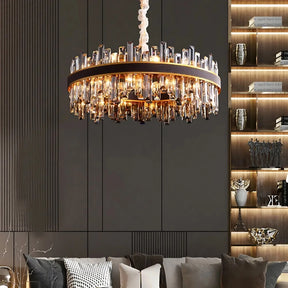 A modern living room features a large, circular Calvi Modern Chandelier by Morsale.com adorned with numerous glass crystals. The dark-paneled wall in the background has built-in shelves displaying various decorations and books. The sofa below is equipped with multiple cushions in neutral tones.