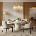 A modern dining room features a circular white table with a white vase holding flowers. The table is surrounded by blue chairs. Above the table hangs a Cadena Chain Tassel Modern Chandelier by Morsale.com with gold accents and cascading fringe details, set against a dark paneled wall.