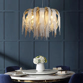 A modern dining room features a circular white table with a white vase holding flowers. The table is surrounded by blue chairs. Above the table hangs a Cadena Chain Tassel Modern Chandelier by Morsale.com with gold accents and cascading fringe details, set against a dark paneled wall.