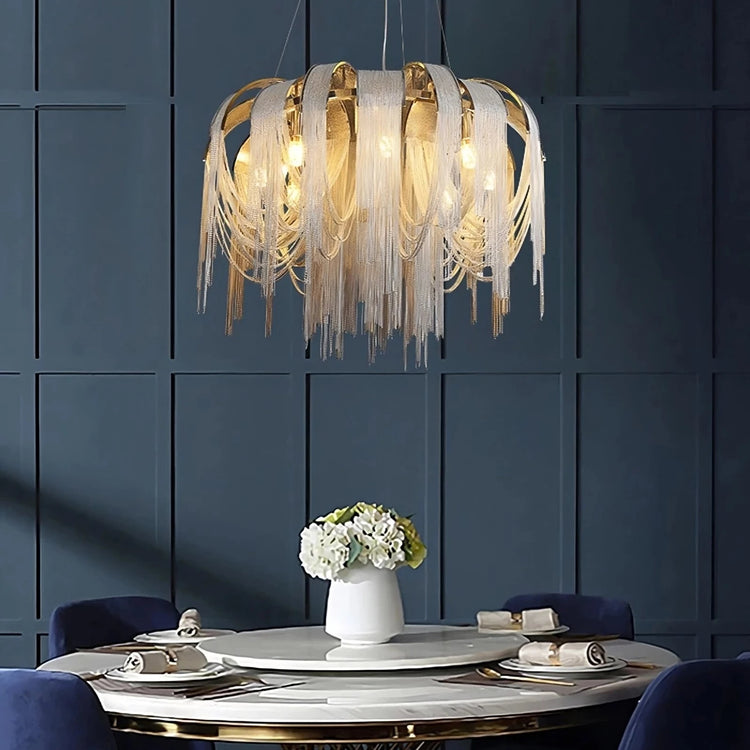 A modern dining room features a circular white table with a white vase holding flowers. The table is surrounded by blue chairs. Above the table hangs a Cadena Chain Tassel Modern Chandelier by Morsale.com with gold accents and cascading fringe details, set against a dark paneled wall.