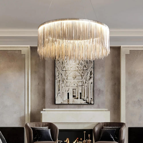The Cadena Modern Chandelier from Morsale.com, a circular LED fixture with cascading metallic fringe, hangs above a fireplace and armchairs in an elegant living room, with black-and-white architectural artwork on the wall behind.