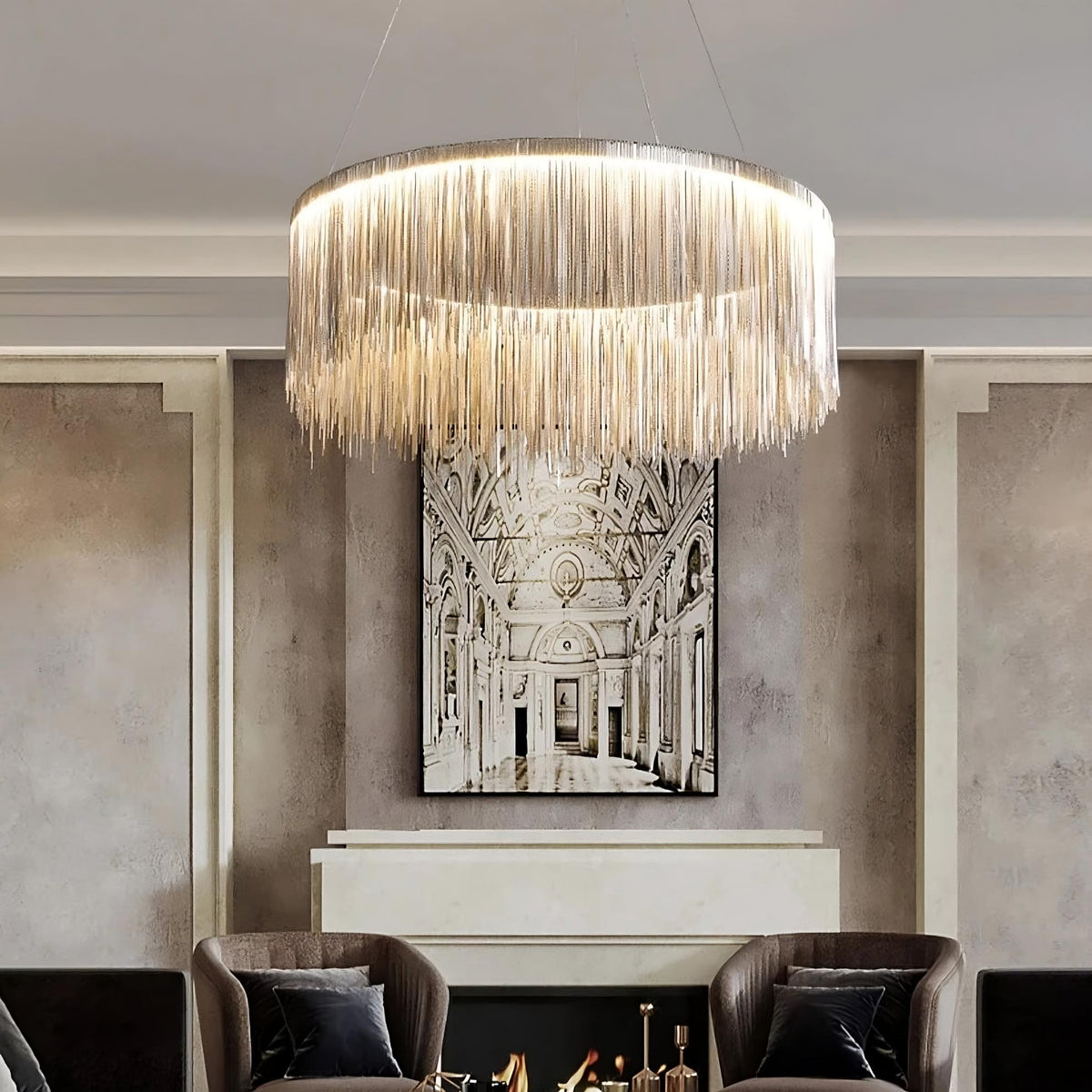 A modern living room features a Morsale.com Cadena Steel Tassel Light Fixture with cascading fringe hanging from the ceiling. Below, energy-efficient LED lighting highlights a fireplace with a black and white architectural painting mounted above it. Two dark armchairs with gray pillows are placed on either side of the fireplace.