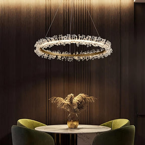 A modern dining area features a round marble table with a vase of pampas grass at its center. Two green velvet chairs are positioned around the table. Above, the Capri Crystal Modern Light Fixture by Morsale.com, adorned with K9 crystals, hangs from the ceiling and warmly illuminates the space.