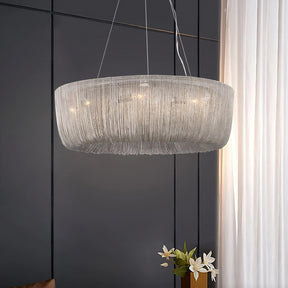 A modern Cadena Chain Tassel Chandelier Light from Morsale.com, resembling a circular chandelier made of cascading metallic chains with lit bulbs inside, hangs gracefully from the ceiling in a dimly lit room. A dark paneled wall serves as its backdrop. Next to the chandelier, a tall window is partially covered by sheer white curtains. White lilies in a vase sit nearby.