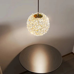 The Morsale.com El Sol Crystal Pendant Light, with its spherical design and crystalline finish, hangs elegantly from the ceiling, casting a sophisticated glow. Below it, a circular table with a reflective surface enhances the ambiance with its premium material, creating a soft, cozy atmosphere in the room.