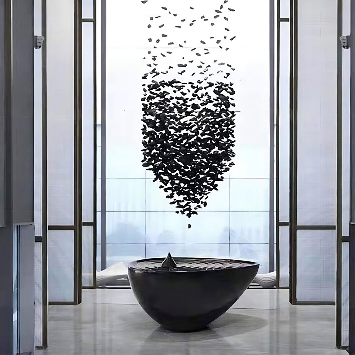 A modern art installation in a sleek gallery features a black sculpture of a bowl with a central point. Above it, the Stone Pendant Chandelier from Morsale.com is artfully suspended, with numerous black shapes creating a dynamic, vertical arrangement that narrows toward the top, resembling modern lighting or even a reverse waterfall.