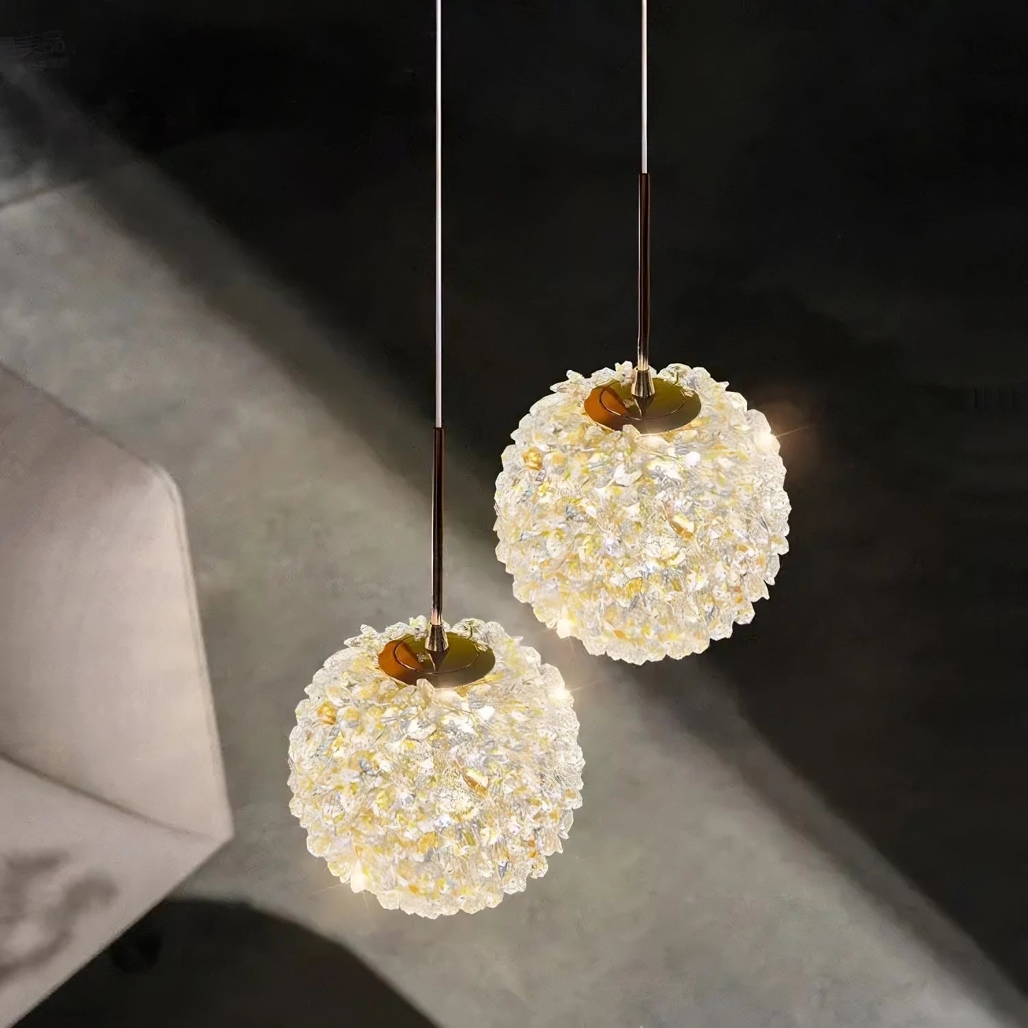 Two spherical hanging lights with a floral clustered design, reminiscent of the El Sol Crystal Pendant Light from Morsale.com, are shown illuminated against a dark background. Each light is suspended by a slender wire and emits a soft, warm glow from premium materials, creating an elegant illumination. An angled view of a gray chair is visible below.