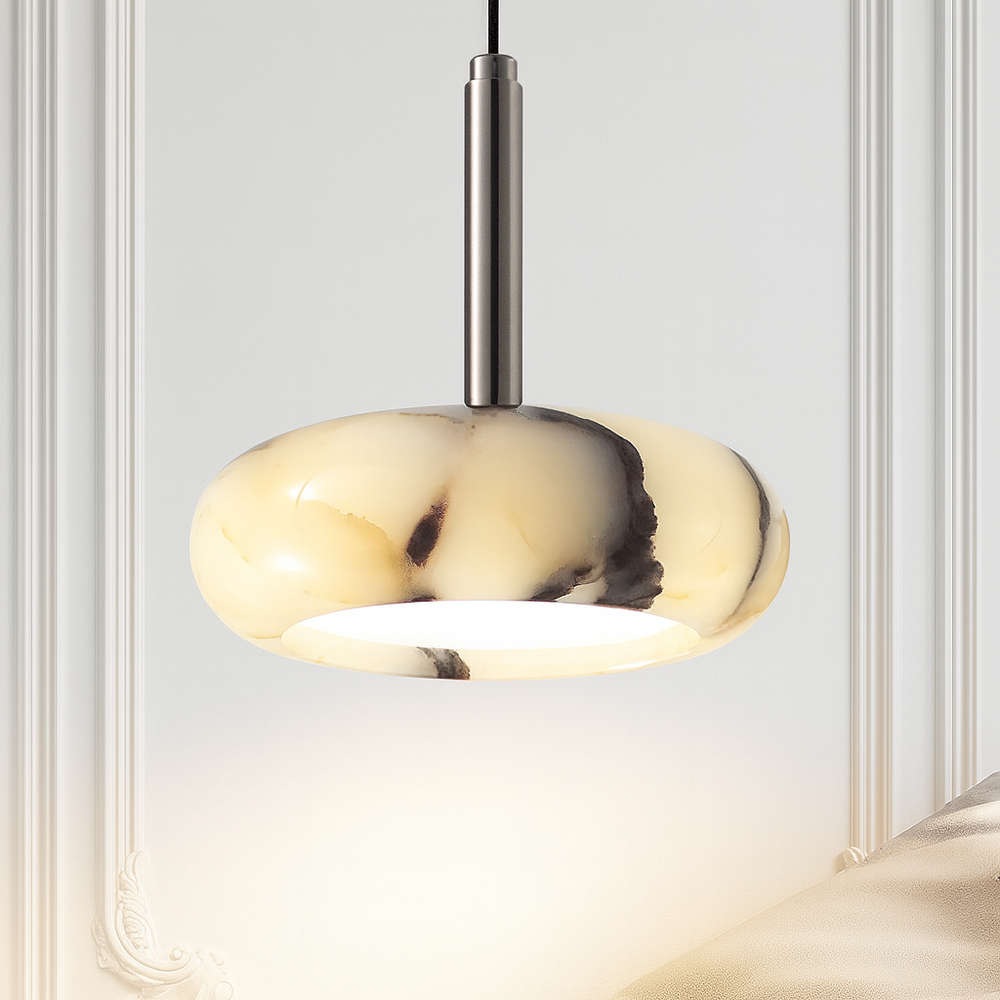 The Luna Natural Marble Pendant by Morsale features a round, marbled cream and black stone shade suspended from a metal fixture. Lit against a white decorative wall, it casts a warm glow and showcases the elegance of natural marble lighting.