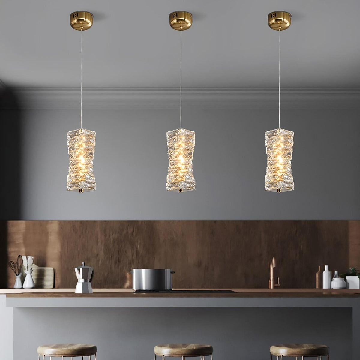 Three elegant Morsale.com Bacci Crystal pendant light fixtures with handmade crystal shades hang above a modern kitchen counter, casting a warm glow on neatly arranged kitchen accessories.