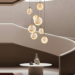 A modern interior design features a spiral staircase with curved beige walls. In the foreground, a round white table adorned with vases and decor items draws the eye. Suspended above are multiple glowing circular lights and the stunning Moonshade Natural Marble Pendant Light Fixture from Morsale.com, creating a warm and elegant atmosphere.