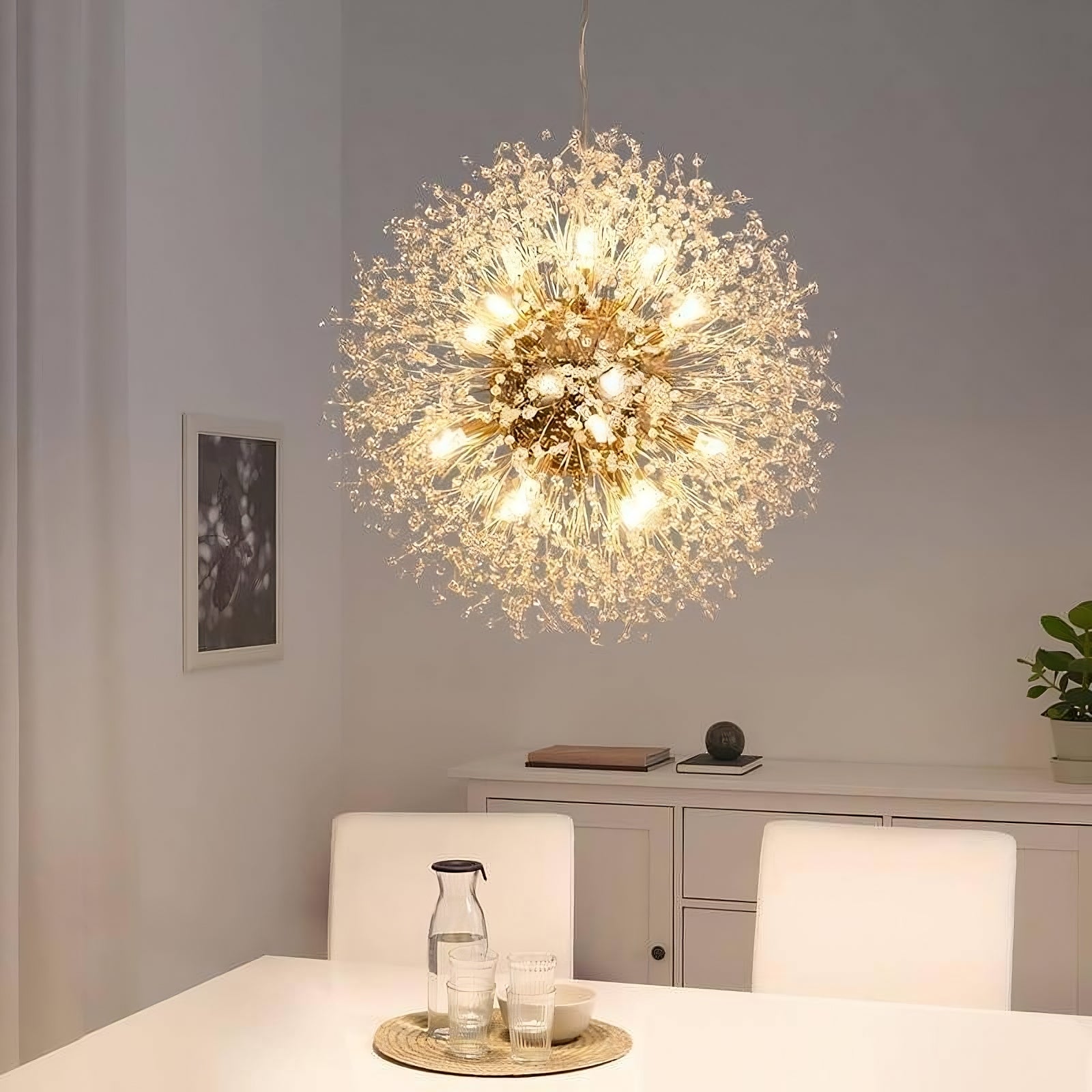 A modern dining room features the El Sol Crystal Dandelion Lighting Fixture from Morsale.com, adorned with K9 Crystals and numerous small LED bulbs, radiating elegance. Below, a set dining table with white chairs stands ready. The background showcases a white sideboard, a small plant, and a framed picture on the wall.