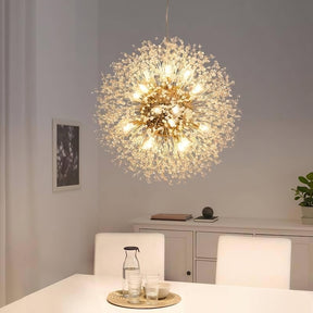 A modern dining room features the El Sol Crystal Dandelion Lighting Fixture from Morsale.com, adorned with K9 Crystals and numerous small LED bulbs, radiating elegance. Below, a set dining table with white chairs stands ready. The background showcases a white sideboard, a small plant, and a framed picture on the wall.