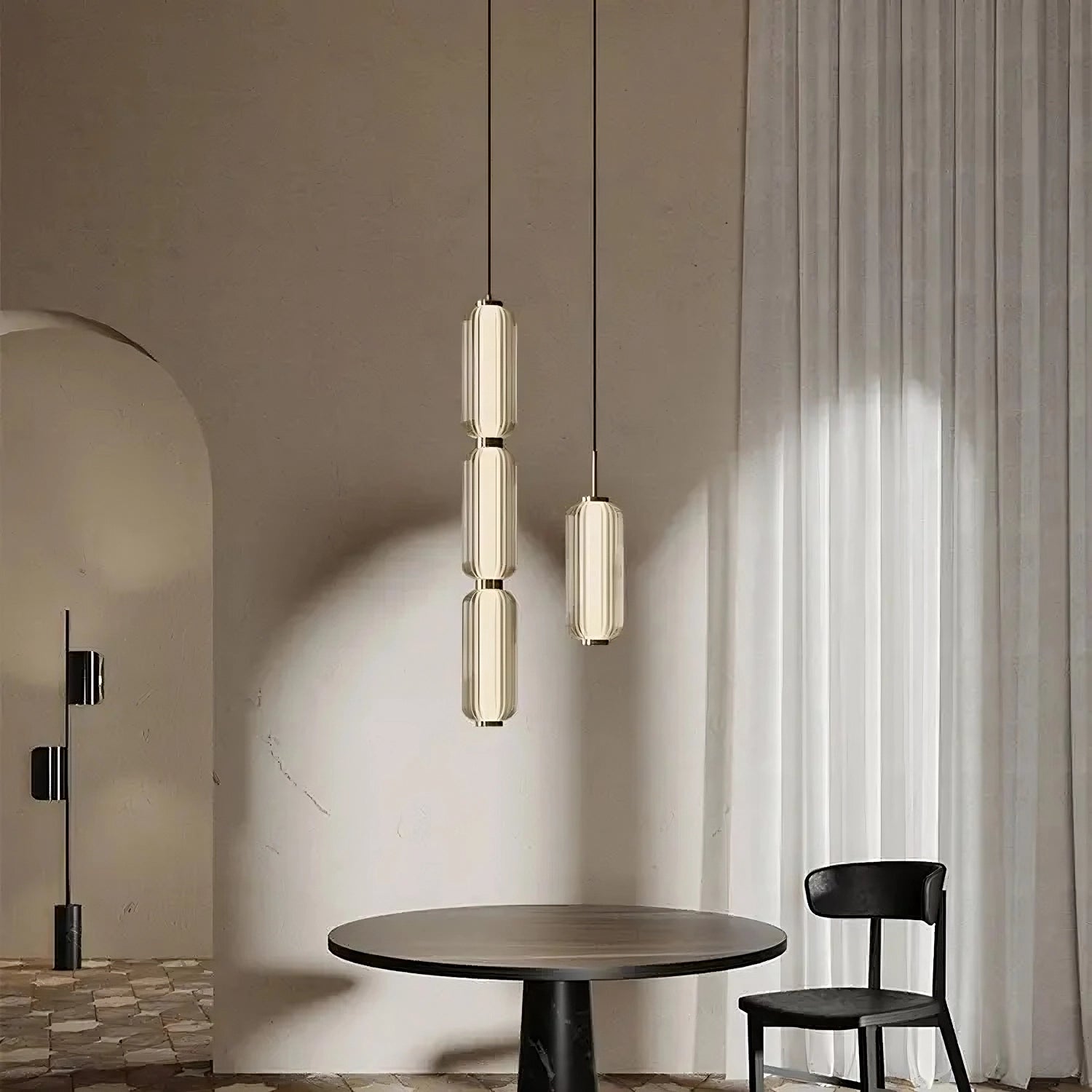 A minimalist room featuring a round, dark wooden table with a black chair beside it. Three elongated Modern Minimalist LED Pendant Light Fixtures from Morsale.com hang from the ceiling, casting illumination. The back wall has an archway and a sheer curtain that allows soft light to filter through.