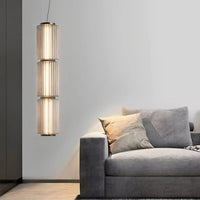 A modern living room with a gray sofa, pillows, a textured rug, and the Luma Modern Glass Pendant by Morsale.com—a vertical cylindrical dimmable LED fixture with three sculpted glass shades—hanging beside the sofa against a light gray wall.